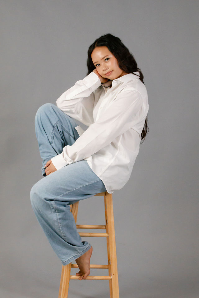 Arianna Luong seated on a wooden stool in a white shirt and jeans